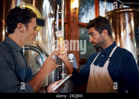 Die Farbe des Biers sagt auch viel darüber aus. Aufnahme von zwei fokussierten jungen männlichen Unternehmern, die tagsüber eine frische Charge Bier in ihrer Bierbrauerei ausprobieren. Stockfoto