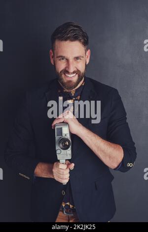 Lächle, du bist offen in der Kamera. Porträt eines trendigen jungen Mannes mit einer Vintage-Kamera vor dunklem Hintergrund. Stockfoto