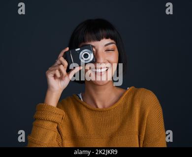 Lächle, du bist vor der Kamera. Studioportrait einer jungen Frau mit Kamera vor dunklem Hintergrund. Stockfoto