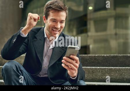 Nur die Nachrichten, die er hören wollte. Ausgeschnittene Aufnahme eines jungen Geschäftsmannes, der eine Faustpumpe macht, während er sein Mobiltelefon ansieht. Stockfoto