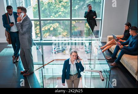 Die Ankunft am Arbeitsplatz für den Tag. Aufnahme einer reifen Geschäftsfrau, die die Treppe hoch geht, während sie in einem geschäftigen Büro auf der Arbeit ein Telefon benutzt. Stockfoto