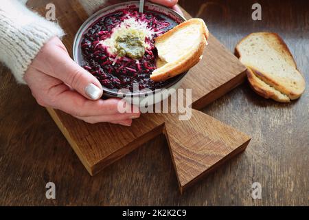 Weibliche Hände halten einen Teller Gemüsesuppe mit Rote Beete, Käse, Sauerrahm und Brot auf arustischem Holzhintergrund Stockfoto