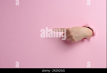 Weibliche Hand ragt aus einem zerrissenen Loch auf einem rosa Papierhintergrund, ein Teil des Körpers zeigt mit dem Zeigefinger zur Seite Stockfoto