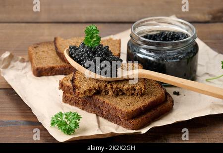 Frischer körniger schwarzer Paddelfisch-Kaviar in braunem Holzlöffel auf einem braunen Tisch, Nahaufnahme Stockfoto