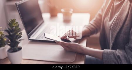 Frau mit Smartphone in der Hand, die im Büro am Laptop arbeitet Stockfoto