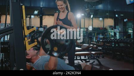 Personal Trainer hilft dem Muskelmann auf der Bank beim Gewichtheben mit der Hakenglocke, die die Muskeln biegt Stockfoto