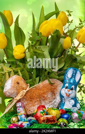 Osterkarten-Vorlage. Ein traditioneller Osterlamm-Kuchen mit Band und einem Osterhasen und vielen anderen Schokoladensüßigkeiten auf gelbem Tulpenstrunk-Hintergrund. Makro. Stockfoto