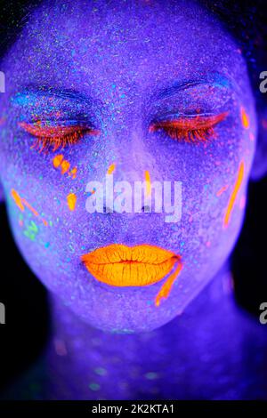 Leuchtende Göttin im Dunkeln. Eine junge Frau posiert mit Neonfarbe auf ihrem Gesicht. Stockfoto