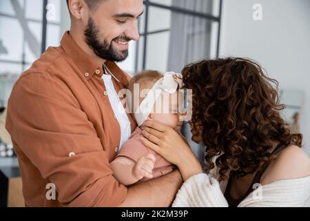 Lockige Mutter berührt Nase von Baby Tochter in Stirnband in der Nähe fröhlichen Mann zu Hause, Stockbild Stockfoto