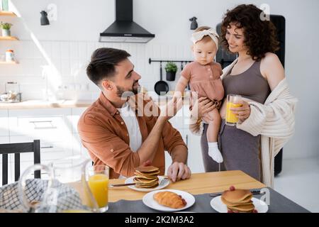 Fröhliche Frau, die ihre kleine Tochter in den Armen hält und dem Ehemann Orangensaft zum Frühstück mitbringt, Stockbild Stockfoto