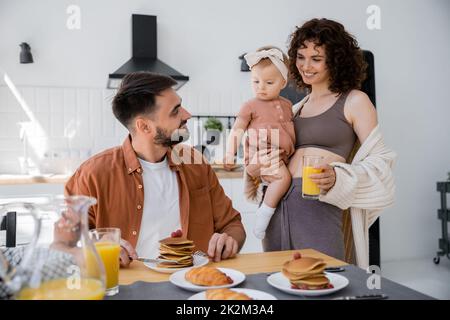 Fröhliche Frau hält in den Armen Baby-Tochter und bringt Orangensaft zum Mann während des Frühstücks, Stockbild Stockfoto