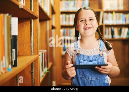 Das ist fast wie ein Buch. Porträt eines fröhlichen jungen Mädchens, das tagsüber auf einem digitalen Tablet unterwegs ist, während es in einer Bibliothek steht. Stockfoto