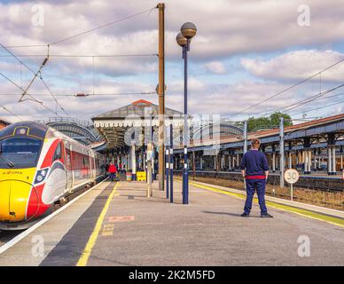 Ein Zug ruht auf einer Plattform mit einem historischen Vordach. Ein Besatzungsmitglied steht auf der Plattform und ein Himmel mit Wolken ist oben. Stockfoto