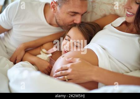 Und bald werden es vier sein. Aufnahme eines glücklichen kleinen Mädchens, das mit ihrem Vater und ihrer Schwangeren im Bett liegt. Stockfoto