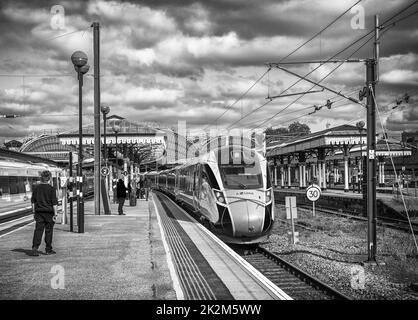 Ein Zug ruht auf einer Plattform mit einem historischen Vordach. Crew-Mitglieder und Passagiere stehen auf dem Bahnsteig. Ein Himmel mit Wolken ist oben. Stockfoto