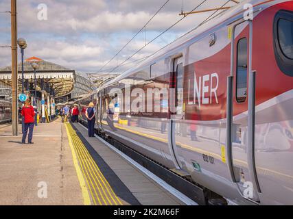 Ein Zug ruht auf einer Plattform mit einem historischen Vordach. Crew-Mitglieder und Passagiere stehen auf dem Bahnsteig. Ein Himmel mit Wolken ist oben. Stockfoto