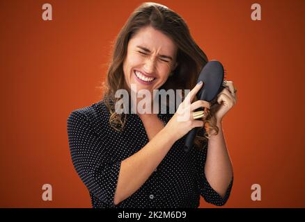 Also... viele... Knoten. Aufnahme einer attraktiven jungen Frau, die im Studio vor einem roten Hintergrund steht und kämpft, um ihr Haar zu putzen. Stockfoto