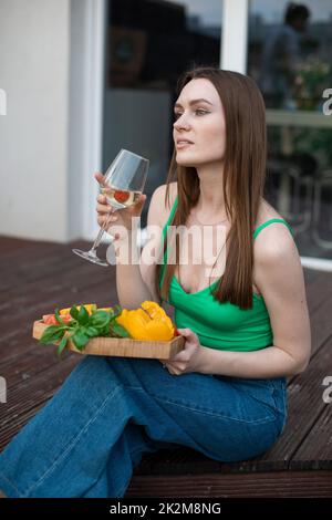 Porträt einer jungen, nachdenklichen, wunderschönen Frau, die auf einer hölzernen Veranda sitzt, ein Glas Wein hält und Gemüse schneidet. Stockfoto