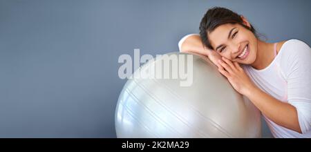 Das Training mit meinem schweizer Ball macht mich glücklich. Eine kurze Aufnahme einer sportlichen jungen Frau, die sich vor grauem Hintergrund auf einen Pilates-Ball lehnt. Stockfoto