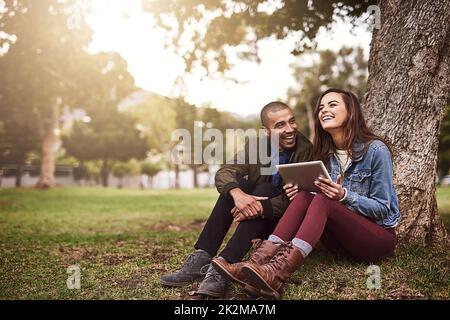 Friedliche Morgen zusammen. Aufnahme eines fröhlichen jungen Paares, das sich mit einem digitalen Tablet draußen in einem Park unter einem Baum sitzend befindet. Stockfoto