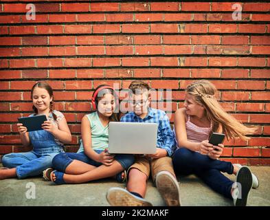 Eine unbeschwerte und miteinander verbundene Kindheit teilen. Aufnahme einer Gruppe von kleinen Kindern, die digitale Geräte gegen eine Ziegelwand verwendeten. Stockfoto