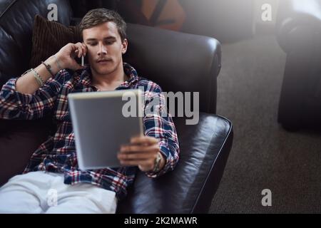 Klar. Ich melde mich gerade an. High-Angle-Aufnahme eines hübschen jungen Mannes, der sein Tablet und Handy benutzt, während er zu Hause auf dem Sofa sitzt. Stockfoto