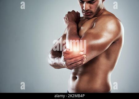 Eine Zeitlang keine Hebegewichte. Ein kurzer Schuss eines sportlichen jungen Mannes, der seinen Ellbogen vor grauem Hintergrund vor Schmerzen hält. Stockfoto