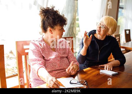 Zwei enthusiastische ältere Frauen tragen legere Kleidung, sitzen am Schreibtisch, diskutieren, machen Notizen in Notizblock im Büro. Stockfoto