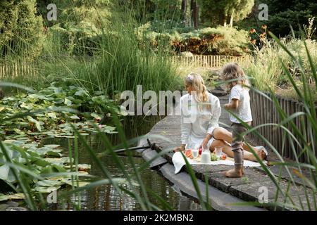 Porträt der Familie, die auf weißem Karli mit Früchten sitzt, Krug mit Milch am Rand des Teiches mit grünen Seerosen in der Nähe des Zauns. Stockfoto