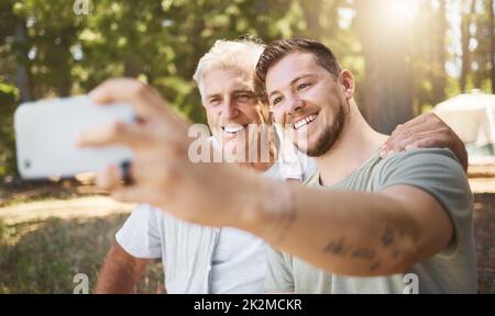 Lächeln. Ausgeschnittene Aufnahme eines hübschen jungen Mannes und seines Vaters, der Selfies beim Camping im Wald macht. Stockfoto