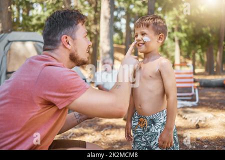 Vergessen Sie nicht Ihre Sonnencreme. Kurzer Schuss eines hübschen jungen Mannes, der seinem entzückenden kleinen Sohn Sonnencreme auf den Platz legt, während er im Wald kampt. Stockfoto