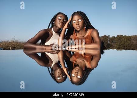 Ungeteilt und immer vereint. Aufnahme von zwei attraktiven jungen Frauen, die neben einem Teich mit ihrer Spiegelung in der Natur sitzen. Stockfoto