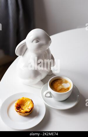 Der Osterhase steht neben einer Tasse Cappuccino und einem Kuchen auf einem Teller, einer weißen Tasse Kaffee. Ostertischdekoration. Speicherplatz kopieren. Stockfoto