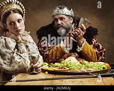 Das königliche Leben kann ermüdend sein. Eine gelangweilte Königin, die bei einem Bankett neben ihrem Mann sitzt. Stockfoto
