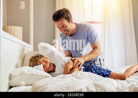 Wach auf, Schlafmütze. Bild eines fröhlichen jungen Mannes, der morgens versucht, seinen Sohn aufzuwecken, indem er zu Hause in einem Bett schläft. Stockfoto