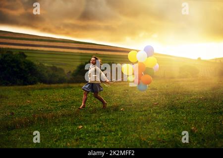 Durch die Felder herumtollen. Aufnahme eines verspielten kleinen Mädchens, das durch ein Feld läuft und dabei einen Haufen Ballons hält. Stockfoto