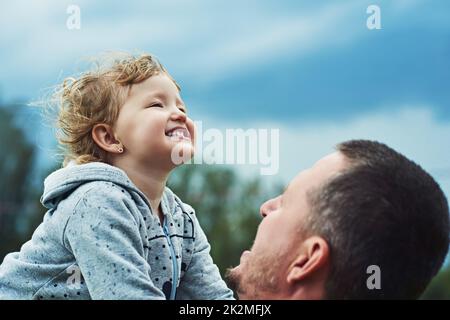 Glücklich wie möglich. Aufnahme eines fröhlichen kleinen Mädchens, das von ihrem Vater an einem bewölkten Tag draußen abgeholt wurde. Stockfoto