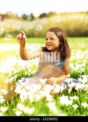 Ein wenig Bubble Spaß in der Sonne. Aufnahme eines entzückenden kleinen Mädchens, das mit Blasen spielt, während es draußen in einem Feld voller Wildblumen sitzt. Stockfoto