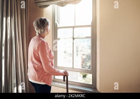 Wann wurden die Tage so grau. Aufnahme einer älteren Frau mit einem Stock, der zu Hause nachdenklich aus dem Fenster schaute. Stockfoto