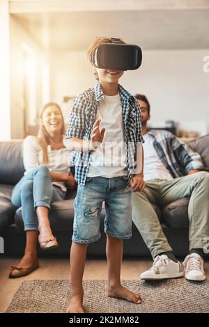 Der Einstieg in eine virtuelle Welt. Aufnahme eines kleinen Jungen, der zu Hause ein Virtual-Reality-Headset verwendet, mit seinen Eltern im Hintergrund. Stockfoto