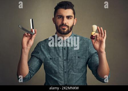 Bart wird gerade gebaut. Studioaufnahme eines hübschen jungen Mannes, der einen Rasiermesser und eine Rasierbürste in den Händen hält. Stockfoto