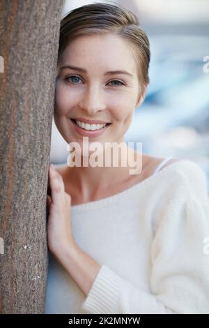 Es gibt so viele schöne Gründe, glücklich zu sein. Porträt einer lächelnden jungen Frau, die sich draußen an einen Baum lehnt. Stockfoto