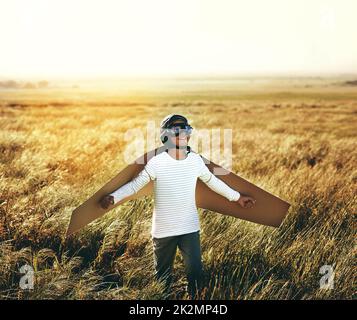 Kinder lieben es, ihre Phantasie fliegen zu lassen. Aufnahme eines Jungen, der vorgibt, mit einem Paar Pappflügeln auf einem offenen Feld zu fliegen. Stockfoto