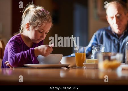 Die Opas haben die Frühstückszeit im Auge. Zugeschnittene Aufnahme eines jungen Mädchens, das am Tisch Getreide isst, mit ihrem Großvater im Hintergrund. Stockfoto