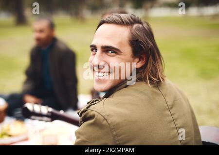 So verbringe ich meine Wochenenden gerne. Porträt eines fröhlichen jungen Mannes, der tagsüber mit seinen Freunden bei einem Picknick draußen saß. Stockfoto