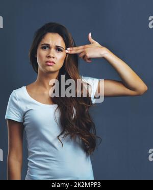 Das Gefühl auf ihrem Verstand. Studioportrait einer jungen Frau, die vor einem dunklen Hintergrund eine Fingergewehrgeste gegen ihren Kopf hält. Stockfoto