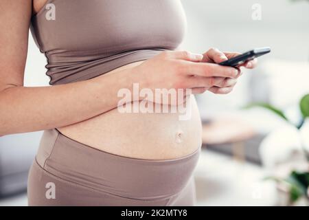 Nahaufnahme des schwangeren weiblichen Bauches. Frau, die eine Smartphone-App in der Wohnung hält und verwendet. Schwangerschaft, Technologie, Online-Shopping, Vorbereitung und Erwartungskonzept. Stockfoto