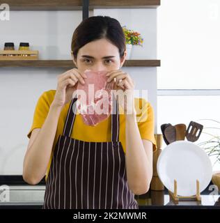 Junge asiatische Hausfrau in einer Schürze gekleidet, die mit beiden Händen ein Stück frisches Steak hält. Stockfoto