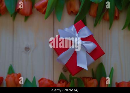 Pinke Tulpen mit grünen Blättern liegen in einer Reihe unten und oben auf einem Holzhintergrund, in der Mitte ist ein Geschenk in einem roten Paket mit einer weißen Schleife. Stockfoto