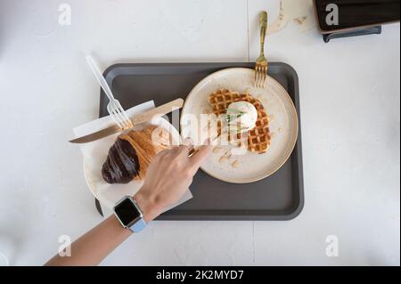Blick von oben auf die Hand mit einem Löffel, der Eiswaffel und Croissant auf dem weißen Tisch im Café ausschaufelt Stockfoto